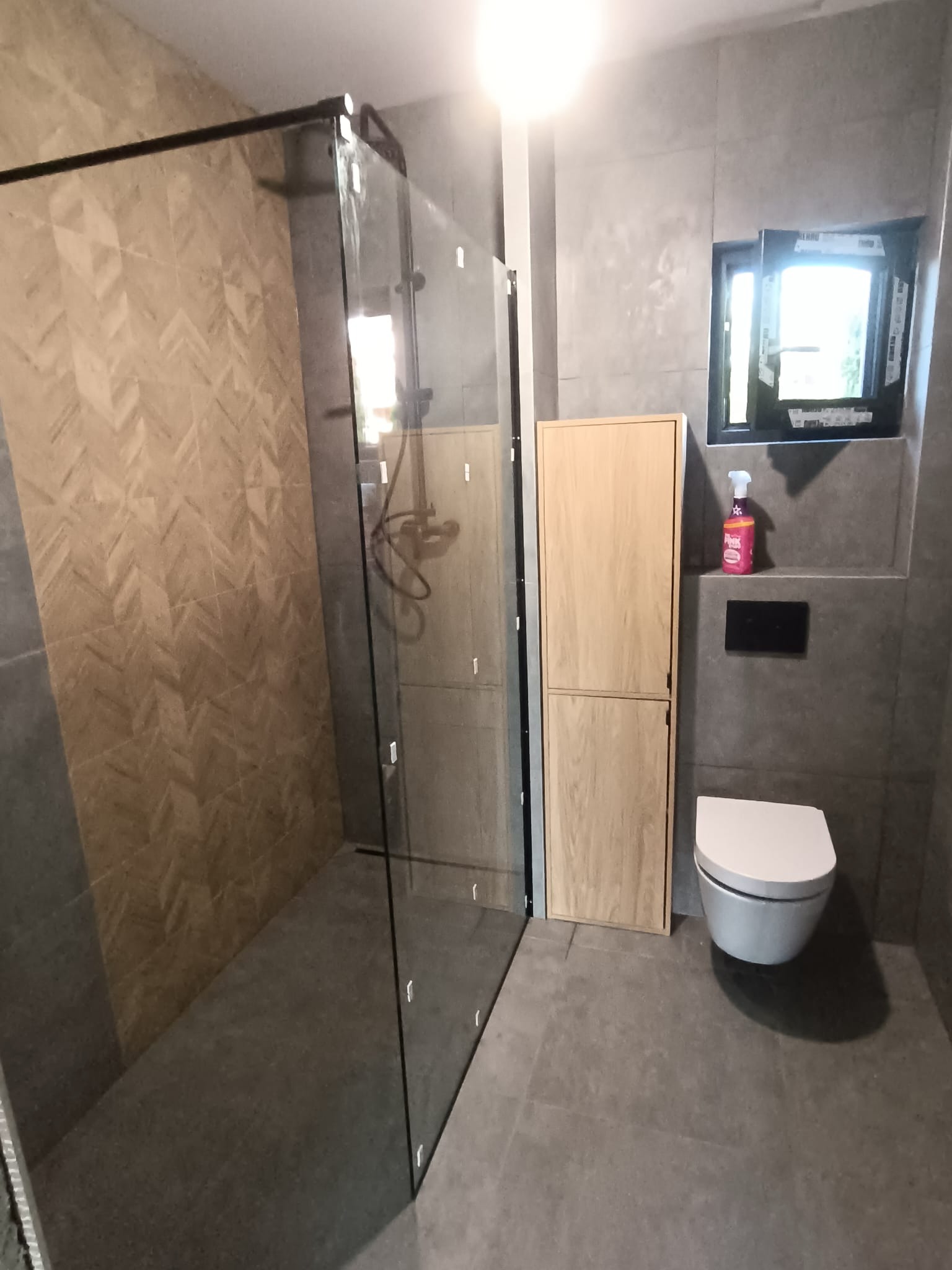 Nowoczesna łazienka z kabiną prysznicową typu walk-in, wykończona szarą płytką i drewnianymi szafkami. Widoczne okno i toaleta podwieszana. Minimalistyczny design.