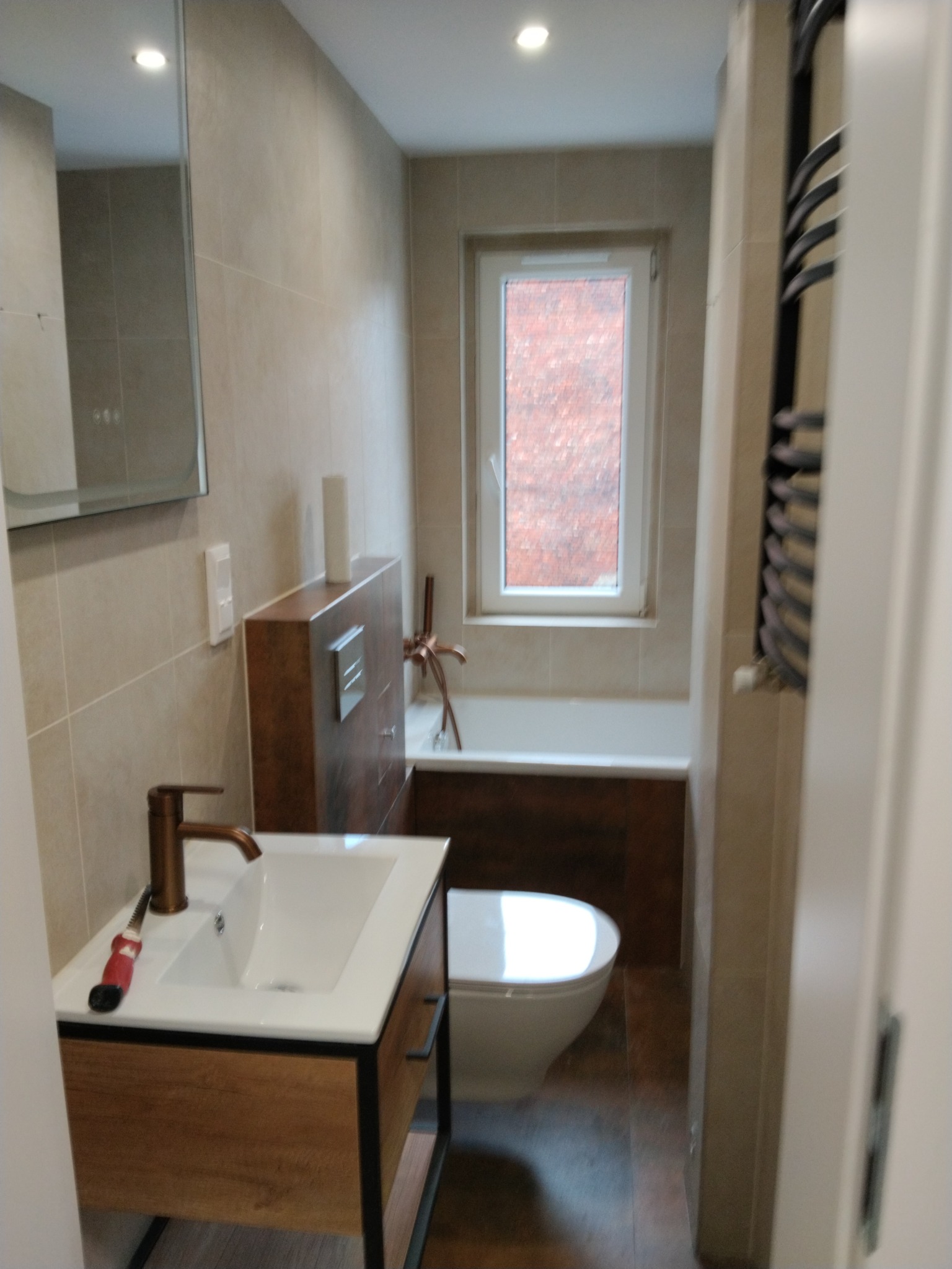 Nowoczesna łazienka z beżowymi płytkami, drewnianą szafką pod umywalką z czarną ramą i baterią w kolorze miedzi. Widoczna toaleta i grzejnik drabinkowy.