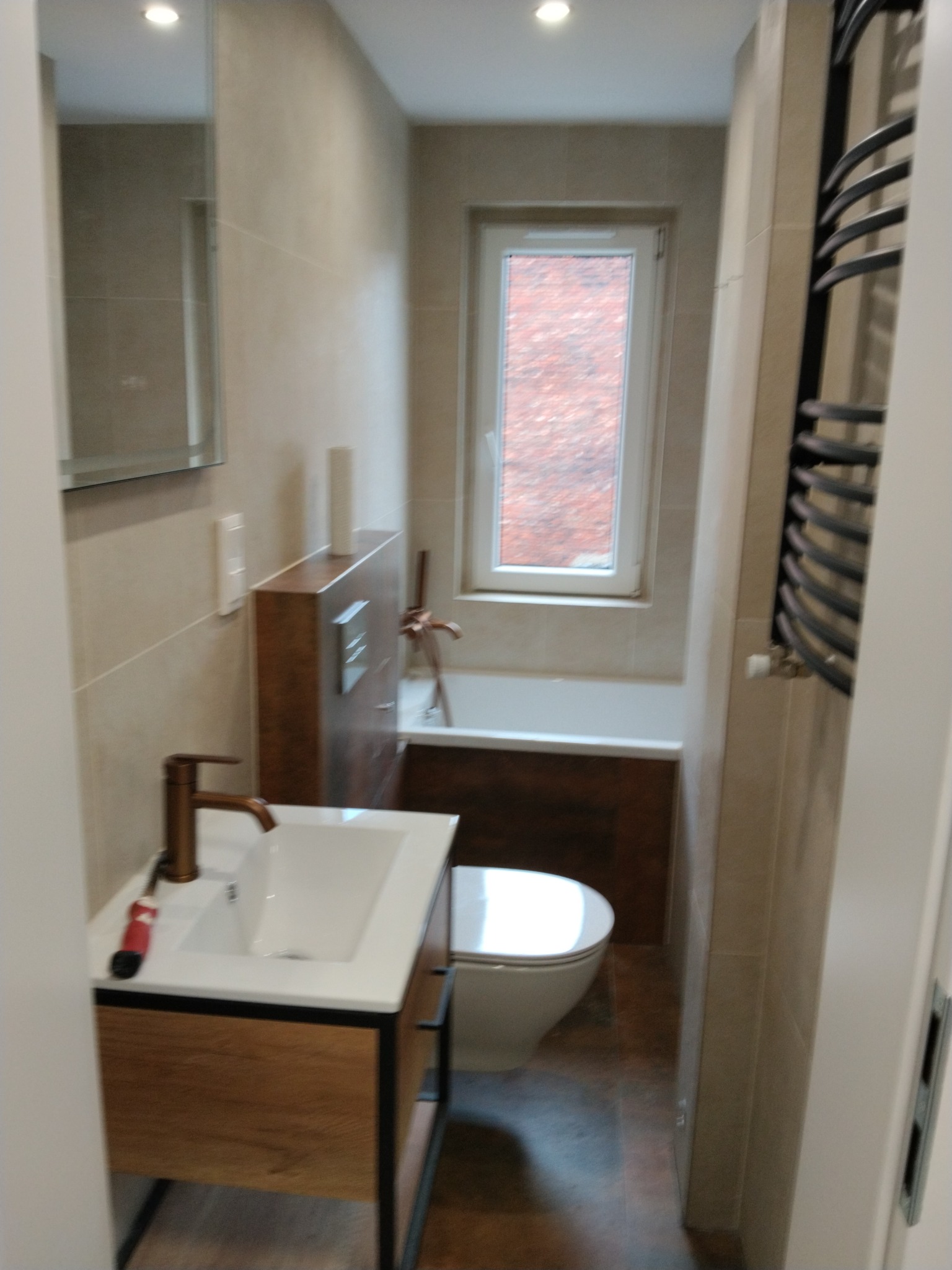 Nowoczesna łazienka z drewnianymi akcentami: szafka pod umywalką, obudowa wanny. Beżowe płytki, grzejnik drabinkowy, okno z widokiem na ceglany budynek.