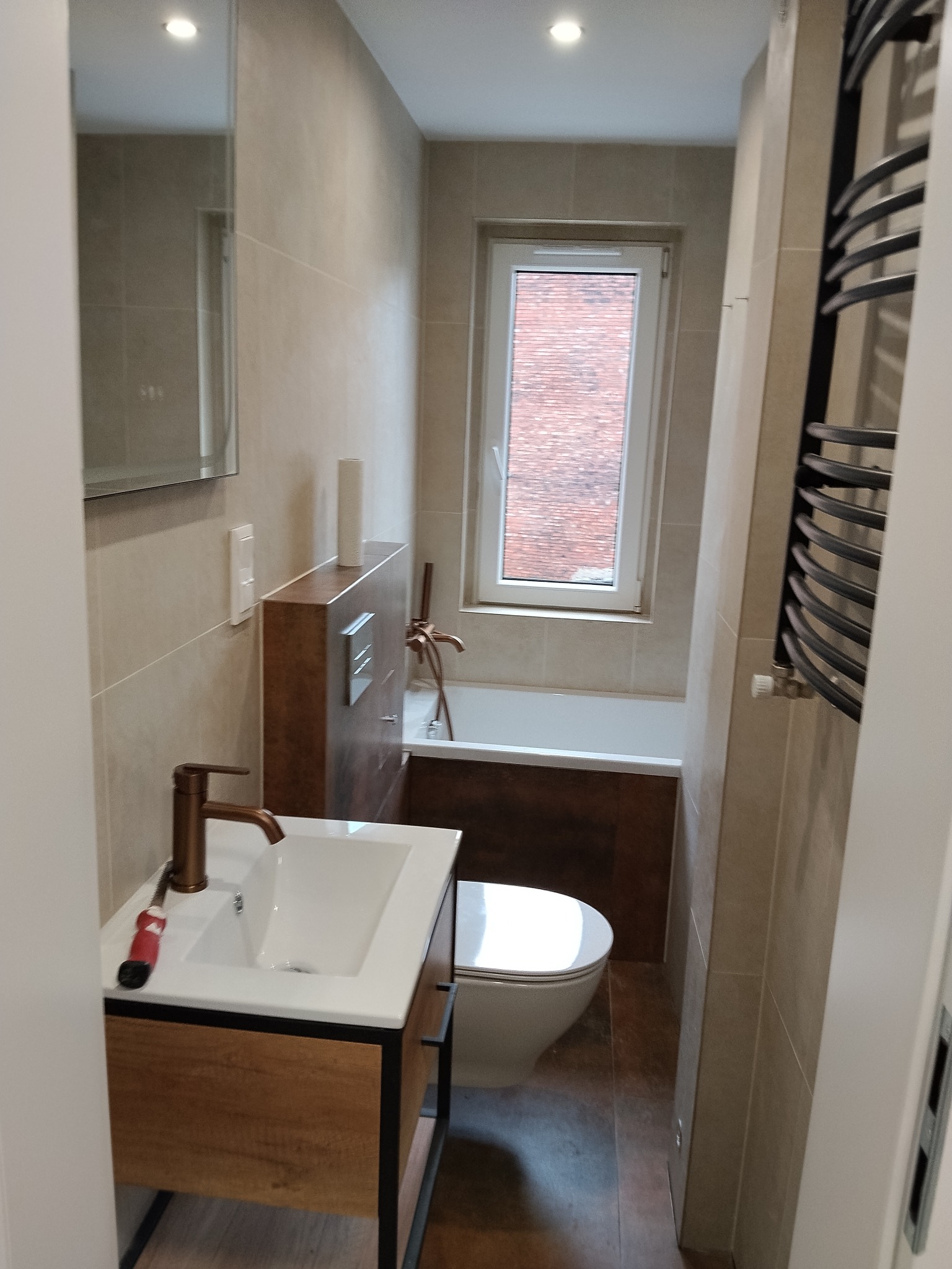 Nowoczesna łazienka z beżowymi płytkami, drewnianą szafką pod umywalką i grzejnikiem drabinkowym. Widok na wannę i okno z widokiem na ceglany budynek.