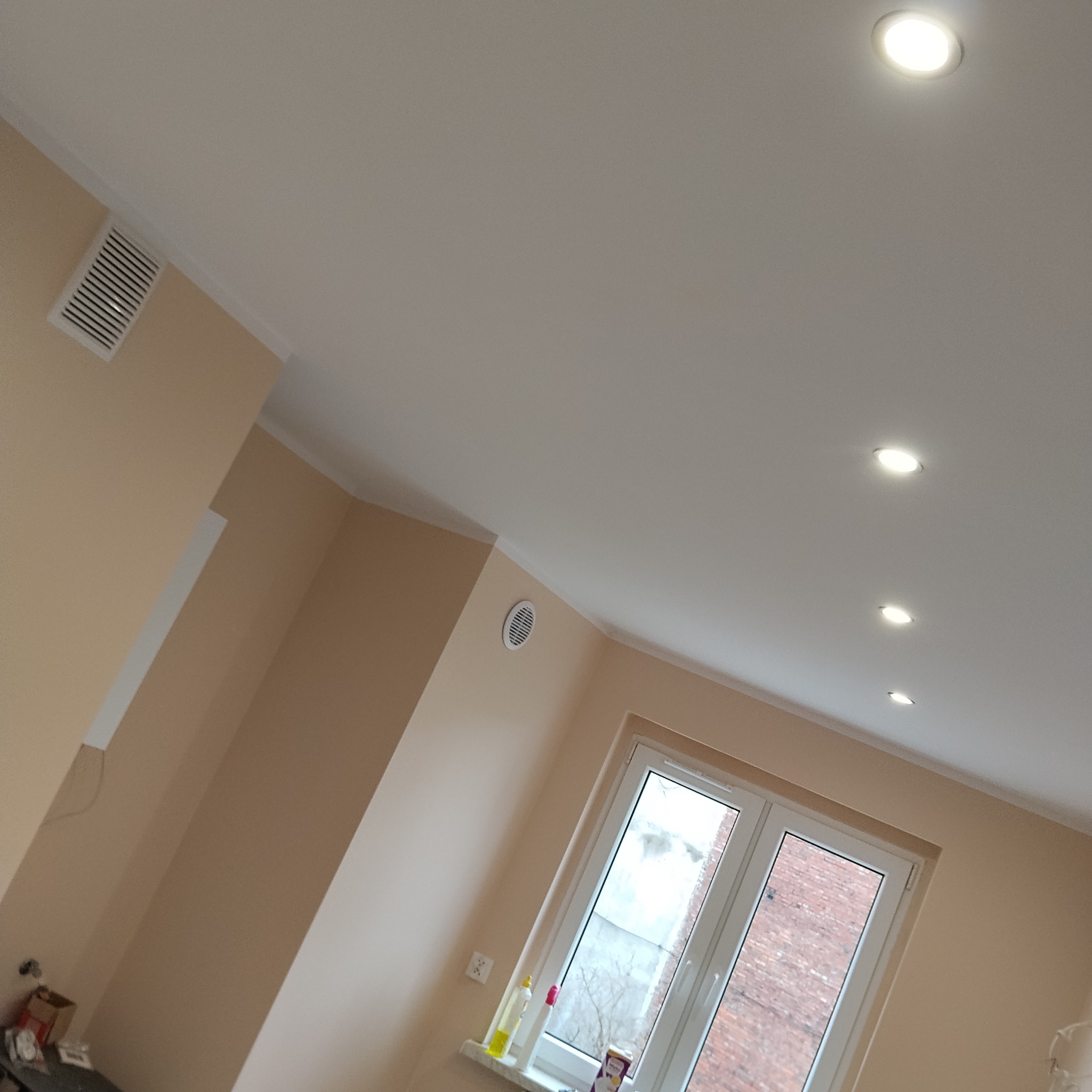 Beżowe ściany i biały sufit z rzędem punktowych lamp LED w pokoju po remoncie. Widoczne okno i kratka wentylacyjna.