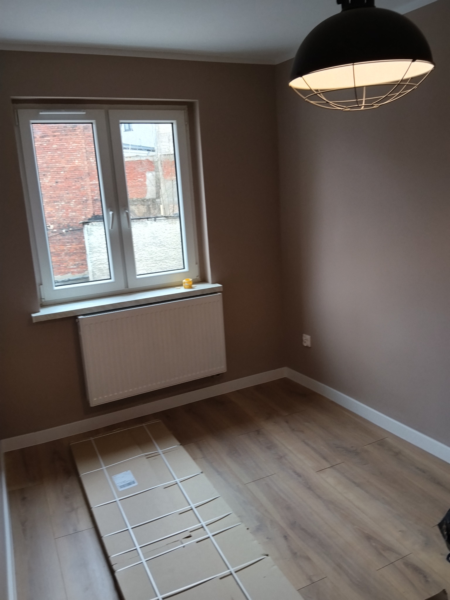 Pokój po remoncie: brązowe ściany, drewniana podłoga, białe okno z widokiem na ceglany budynek. Na podłodze leżą panele w opakowaniu, a z sufitu zwisa nowoczesna lampa.