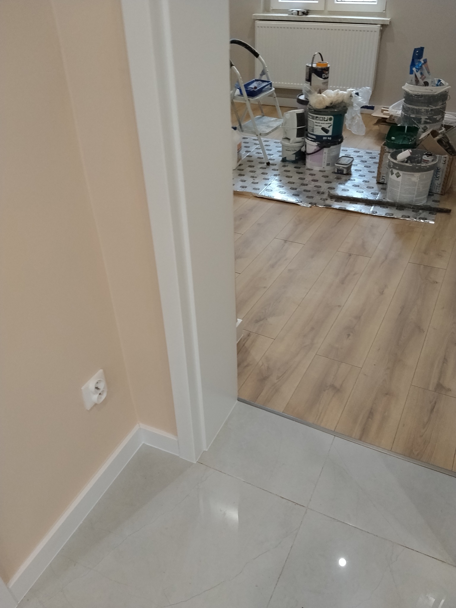 Widok na podłogę z panelami i fragment ściany z gładzią, widoczne puszki z farbą i narzędzia malarskie. Podłoga wyłożona folią ochronną. Ujęcie zza futryny.