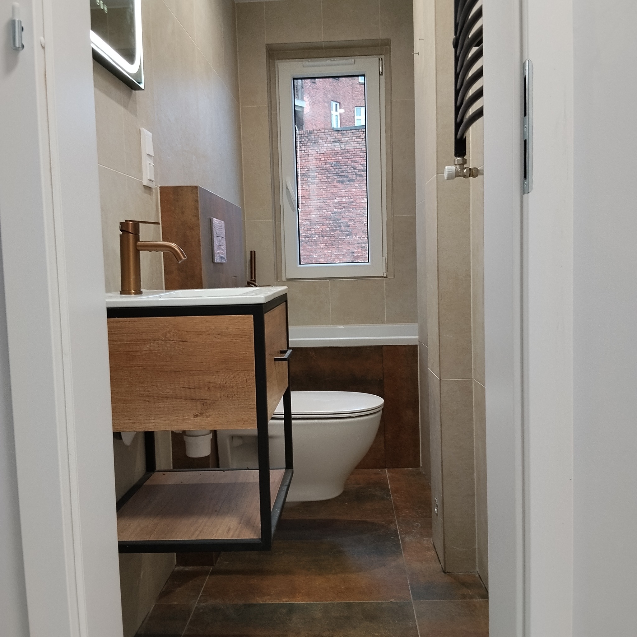 Nowoczesna łazienka z drewnianą szafką pod umywalką i złotą armaturą. W tle okno z widokiem na ceglany budynek. Brązowe płytki podłogowe i beżowe ścienne.