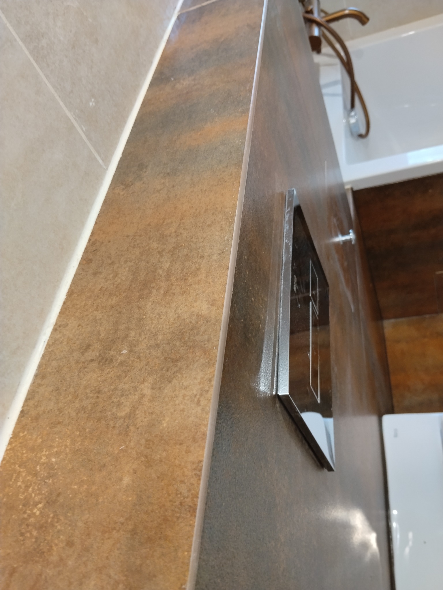 Błyszcząca, brązowa płytka ścienna w łazience z chromowanym przyciskiem spłukującym i fragmentem białej wanny w tle. Ujęcie z bliska, podkreślające fakturę i detale wykończenia.