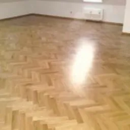 Wypolerowana podłoga drewniana ułożona w jodełkę, widoczne refleksy światła, białe ściany i grzejnik w tle.