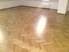 Wypolerowana podłoga drewniana ułożona w jodełkę, widoczne refleksy światła, białe ściany i grzejnik w tle.