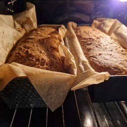 Kuźnia Imprez MAGDALENA MIKOŁAJCZYK - Dwa bochenki domowego chleba w foremkach, wyłożonych papierem do pieczenia, w piekarniku. Widoczne pęknięcia na powierzchni chleba.