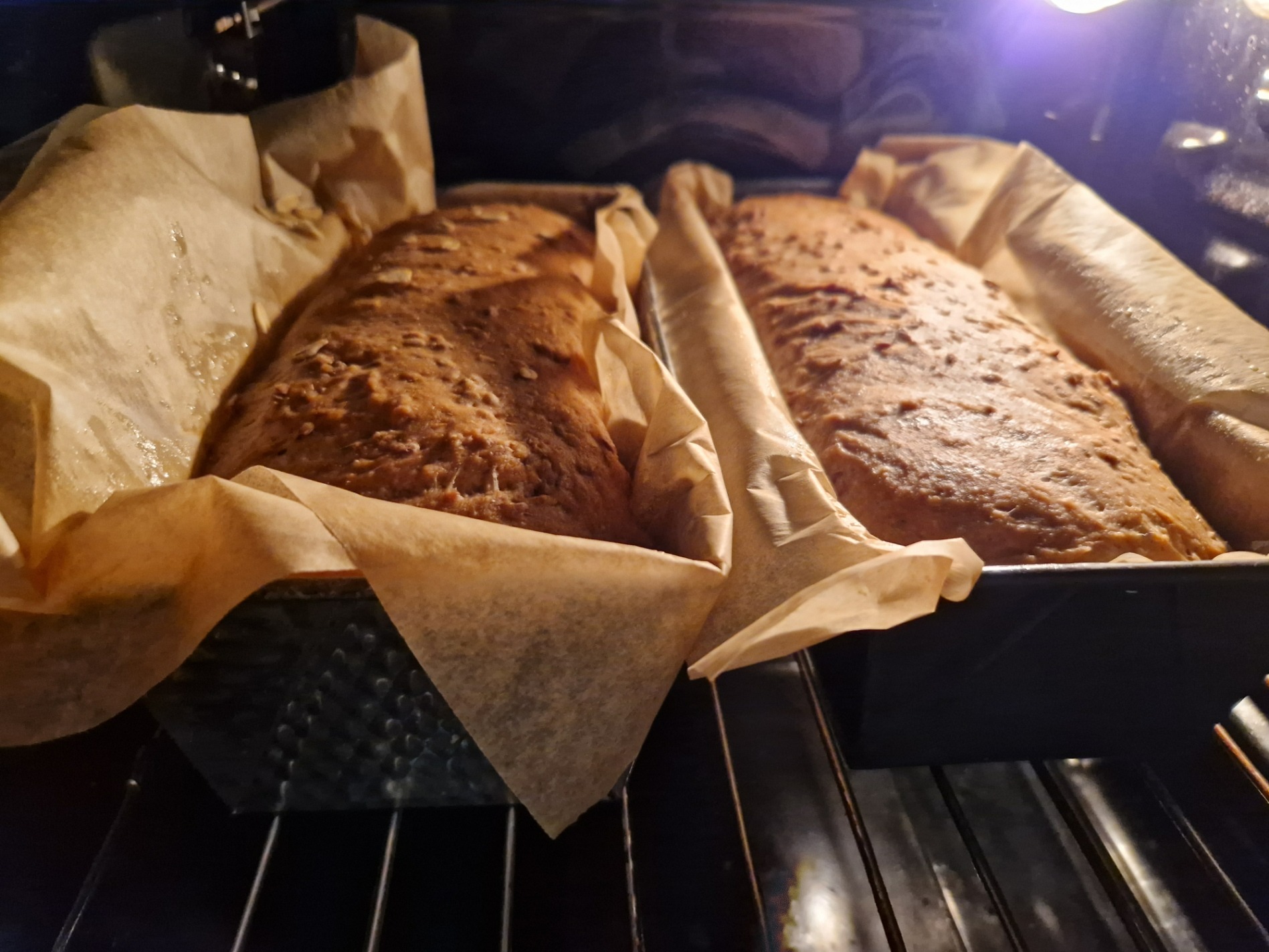 Dwa bochenki domowego chleba w foremkach, wyłożonych papierem do pieczenia, w piekarniku. Widoczne pęknięcia na powierzchni chleba.