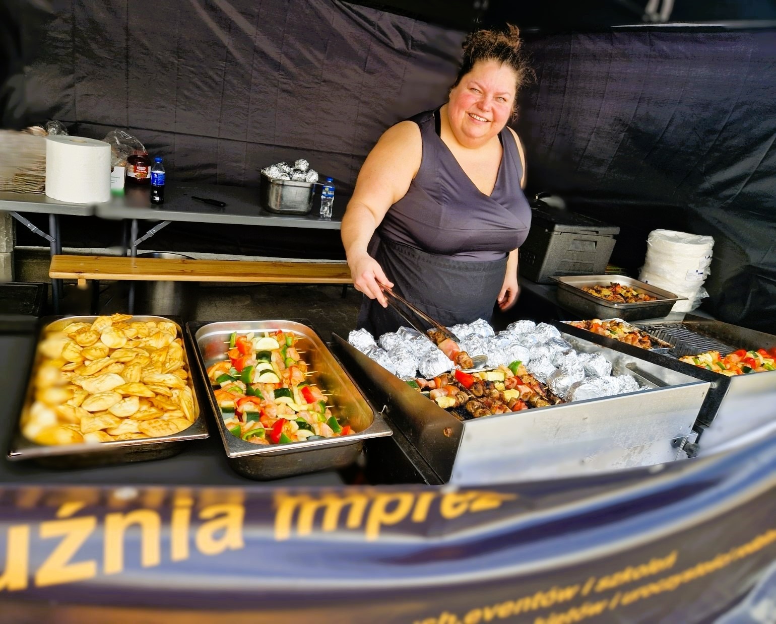Uśmiechnięta kobieta w czarnym fartuchu przygotowuje szaszłyki na grillu, obok tacki z pierogami i warzywnymi szaszłykami, w tle czarne zasłony.