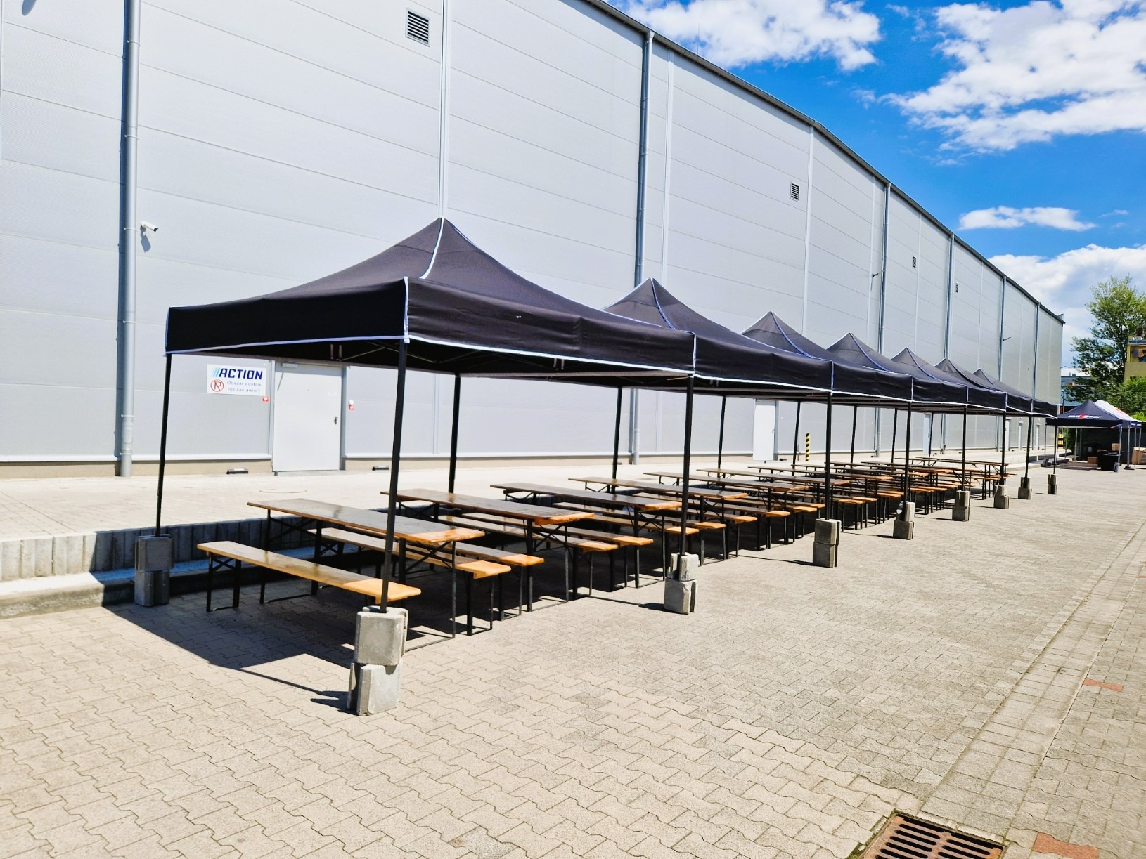 Ustawione rzędy drewnianych stołów i ławek pod czarnymi namiotami na zewnątrz budynku. Przygotowania do eventu.