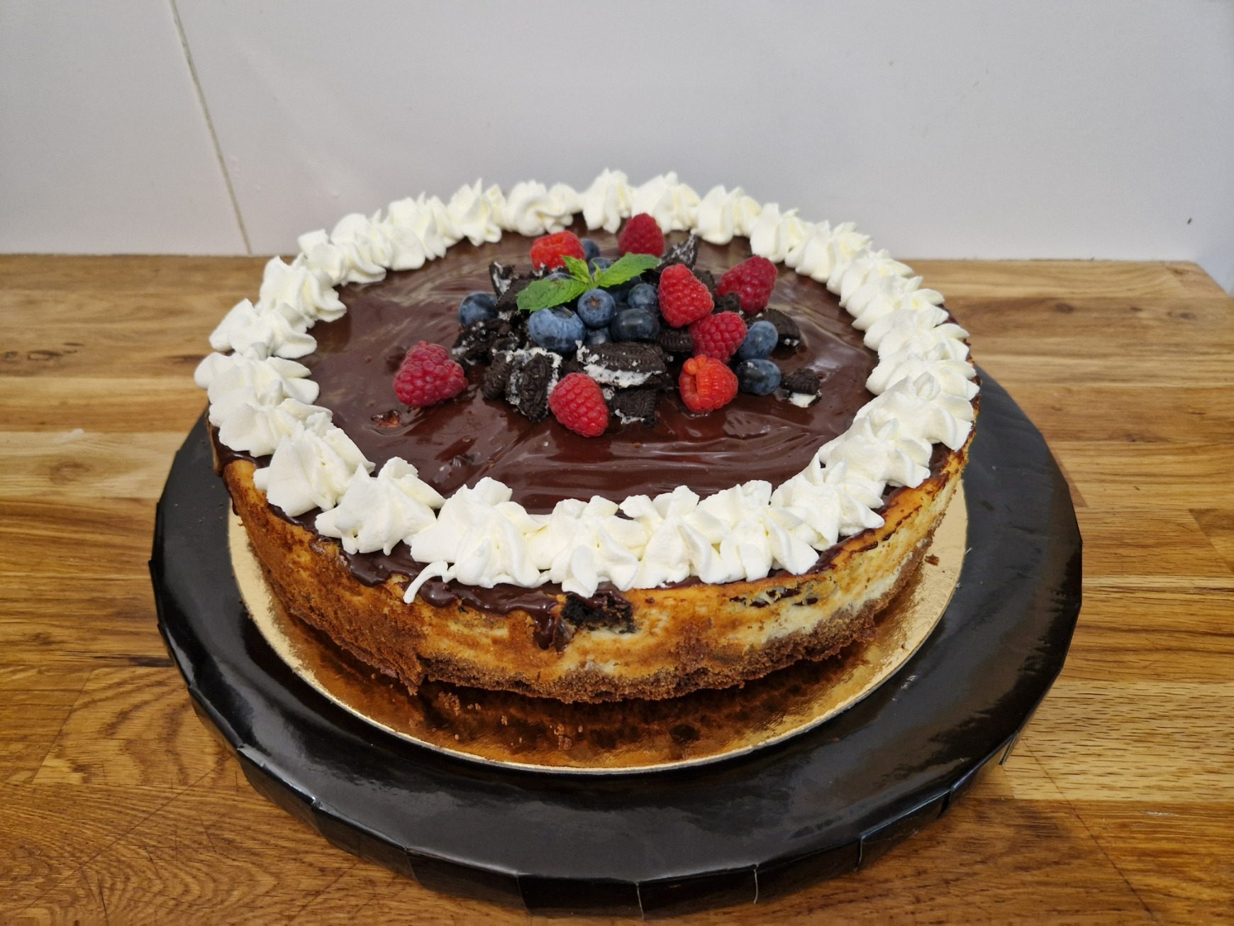 Apetyczny sernik z polewą czekoladową, świeżymi malinami, borówkami i bitą śmietaną na czarnym talerzu, prezentowany na drewnianym blacie. Słodka kompozycja.