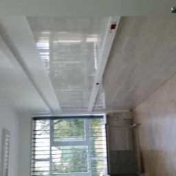 Wykończony sufit z płytek z delikatnym wzorem, widok na okno z żaluzjami i fragment ściany z dekoracyjnymi listwami.