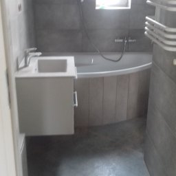 Uslugi remontowo budowlane - Wykończona łazienka w odcieniach szarości z wanną obudowaną płytkami imitującymi drewno, umywalką z szafką i grzejnikiem drabinkowym.