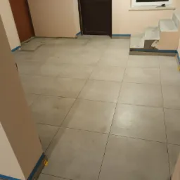 Układanie jasnych płytek podłogowych w korytarzu, widoczne żółte kliny poziomujące i niebieska taśma zabezpieczająca przy ścianach oraz schodach.