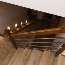 Uslugi remontowo budowlane - Schody z drewnopodobnymi panelami i metalową balustradą, oświetlone punktowymi lampkami w ścianie, widok z góry.