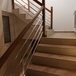 Uslugi remontowo budowlane - Schody wykończone płytkami ceramicznymi z drewnianą balustradą i metalowymi prętami, oświetlenie punktowe na dolnych stopniach, widok z dołu.