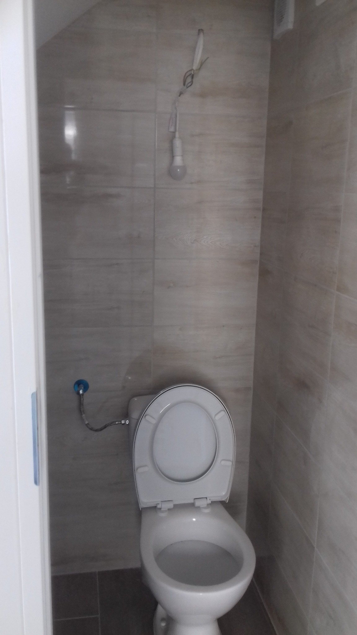 Toaleta po remoncie w małej łazience, widoczne płytki imitujące drewno na ścianach, podłączenie wody do spłuczki i wisząca żarówka na kablu.