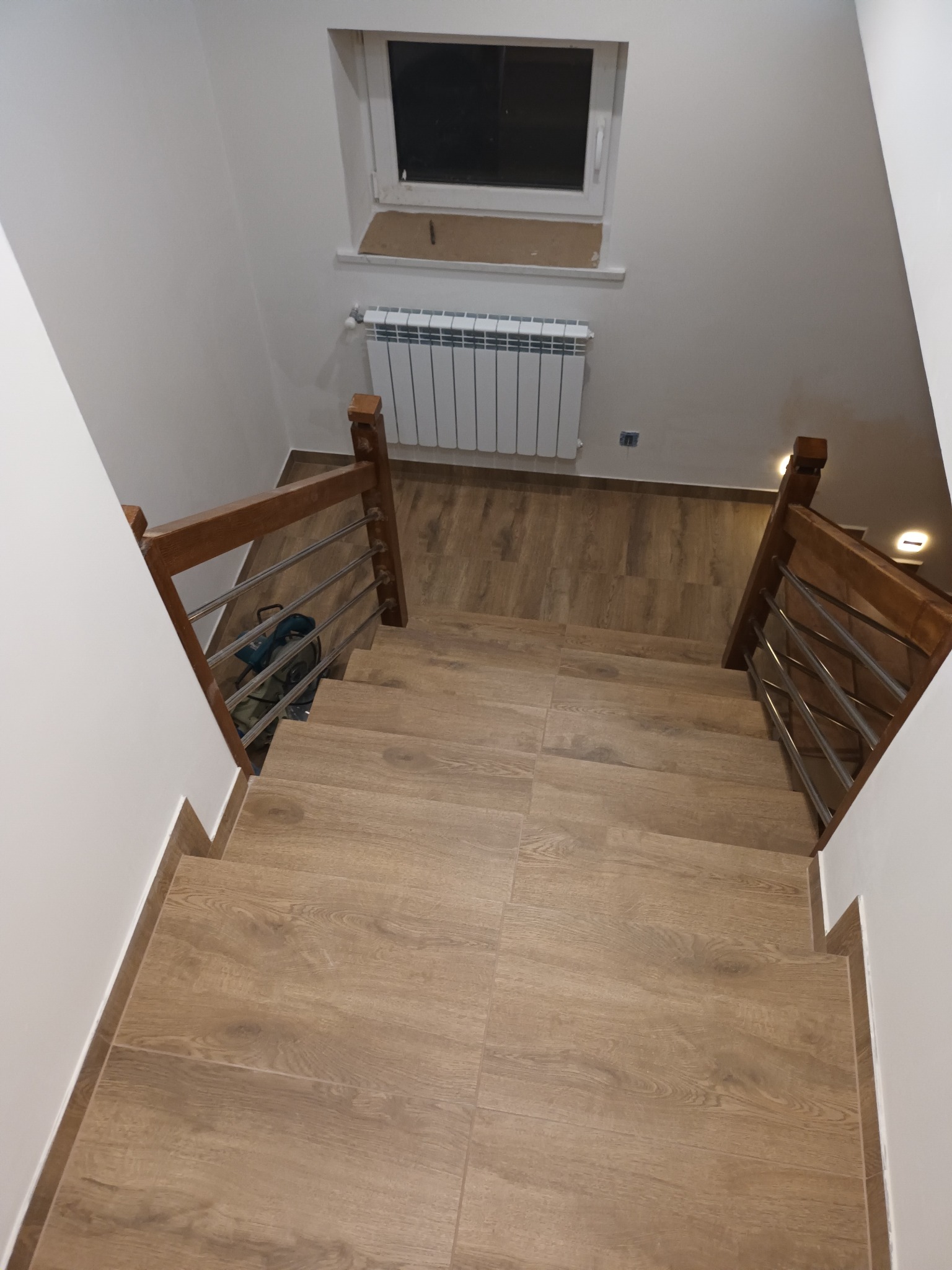 Schody wyłożone płytkami imitującymi drewno, z drewnianą balustradą i metalowymi prętami, widok z góry, białe ściany i grzejnik pod oknem.