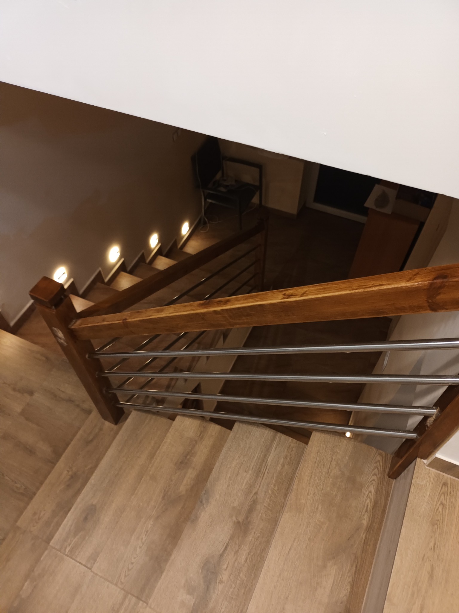 Schody z drewnopodobnymi panelami i metalową balustradą, oświetlone punktowymi lampkami w ścianie, widok z góry.