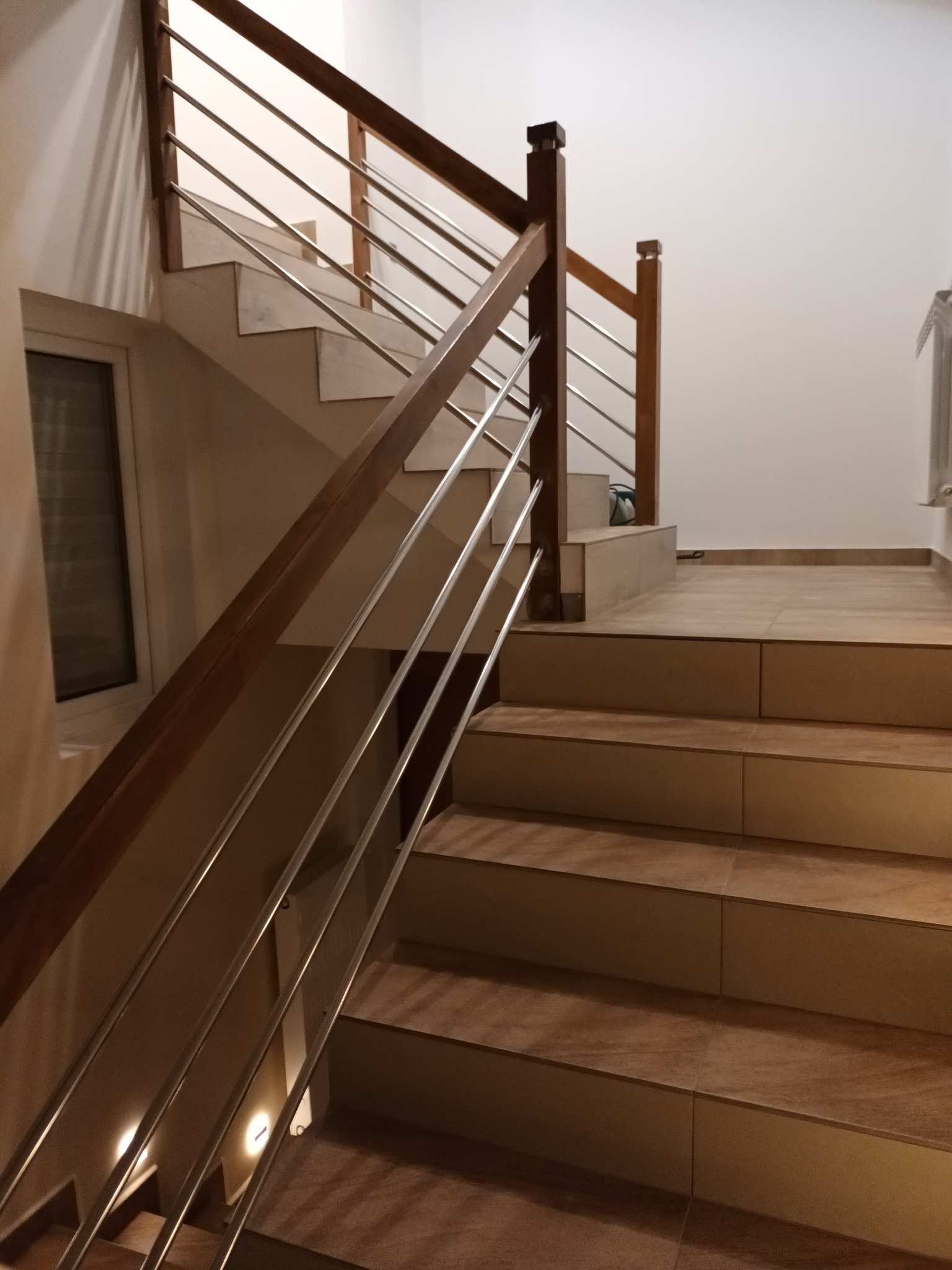 Schody wykończone płytkami ceramicznymi z drewnianą balustradą i metalowymi prętami, oświetlenie punktowe na dolnych stopniach, widok z dołu.