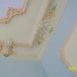 Dekoracyjny sufit z gipsowymi ornamentami w pastelowych kolorach, widoczne detale sztukaterii z motywami roślinnymi i geometrycznymi.