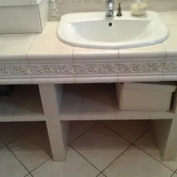 Umywalka w łazience z kremowymi płytkami i zdobieniami, szafka z półkami, pudełka do przechowywania. Ujęcie z bliska na detale wykończenia.