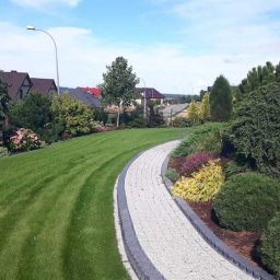 OGRODY PILARSCY mamy zielone pojecie..... - Utrzymany w idealnym stanie trawnik ze ścieżką z jasnej kostki brukowej z ciemnymi obrzeżami, prowadzącą przez zadbane nasadzenia krzewów i kwiatów, na tle zabudowy jednorodzinnej.
