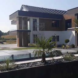 OGRODY PILARSCY mamy zielone pojecie..... - Nowoczesny ogród z palmami i zadaszonym basenem na tle dwukondygnacyjnego domu w słoneczny dzień.