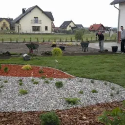 Układanie trawy z rolki w ogrodzie z wydzielonymi strefami: trawnik, kora, kamień; widoczne narzędzia ogrodnicze i pracownicy podczas realizacji.