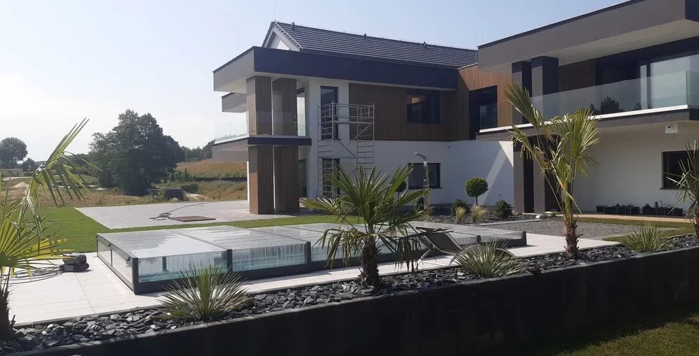 Nowoczesny ogród z palmami i zadaszonym basenem na tle dwukondygnacyjnego domu w słoneczny dzień.