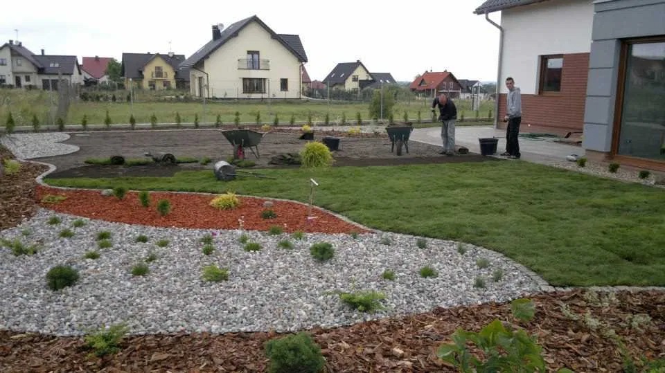 Układanie trawy z rolki w ogrodzie z wydzielonymi strefami: trawnik, kora, kamień; widoczne narzędzia ogrodnicze i pracownicy podczas realizacji.