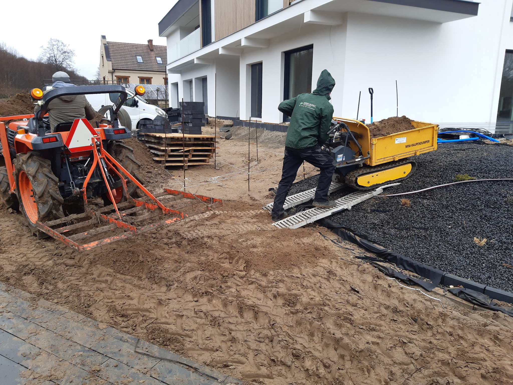 Przygotowanie terenu pod ogród: traktor z broną wyrównuje ziemię, a pracownik wywozi ziemię mini-wywrotką na gąsienicach, widoczne obrzeża i drenaż z kamienia.