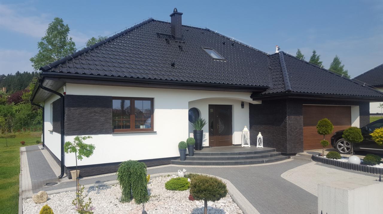 Nowoczesny dom jednorodzinny z czarną dachówką i elementami elewacji wykończonymi ciemnym kamieniem, otoczony zadbanym ogrodem z ozdobnymi krzewami i ścieżką z kostki brukowej.