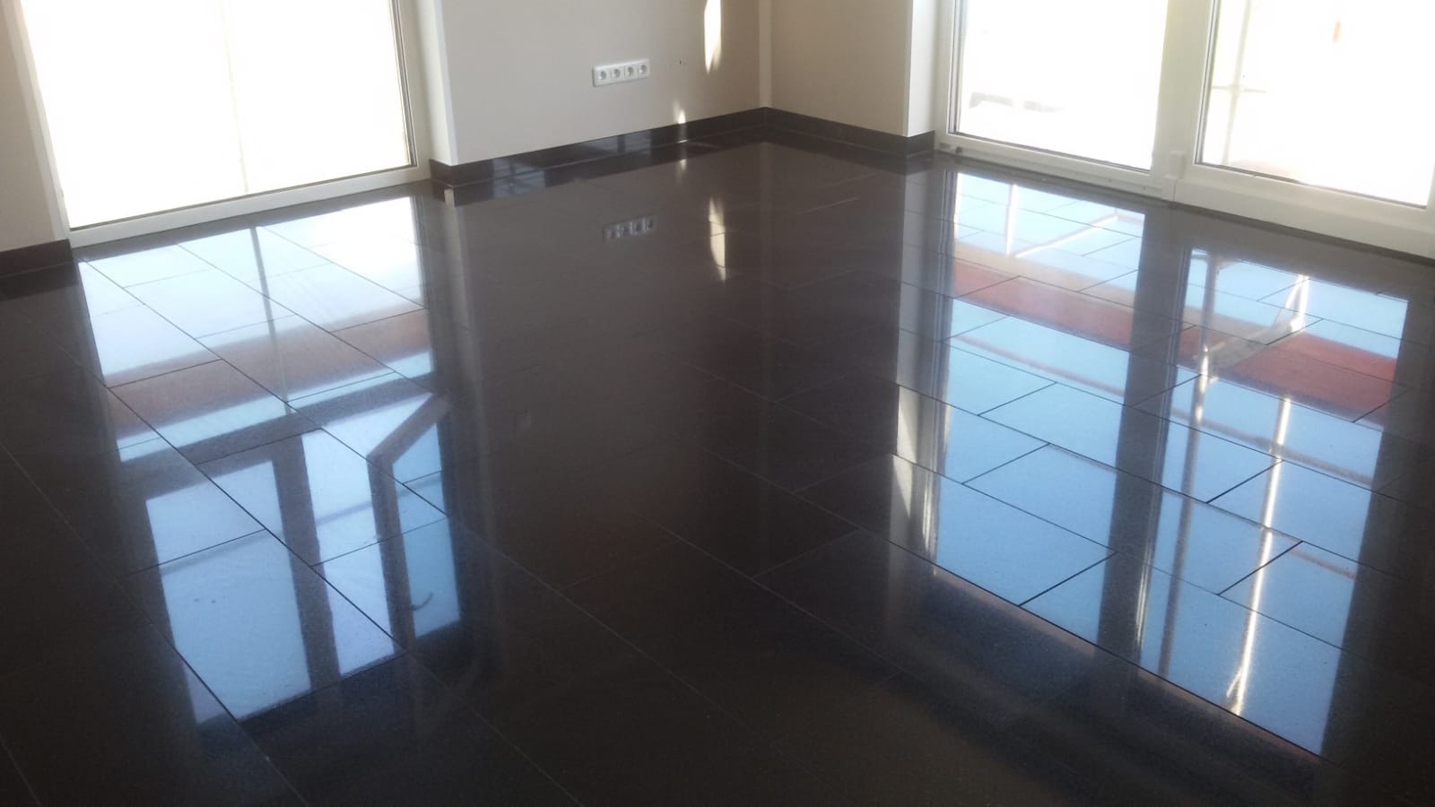 Błyszcząca, ciemna podłoga z płytek ceramicznych w nowym, pustym pokoju z dużymi oknami balkonowymi, odbijającymi światło dzienne.