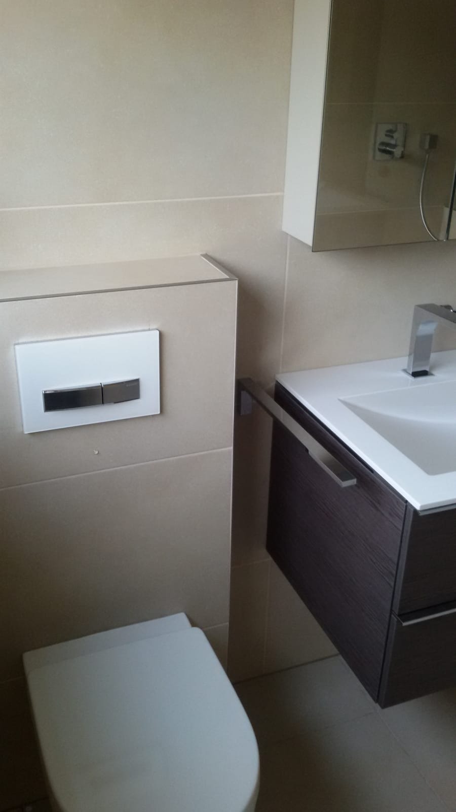 Nowoczesna łazienka z podwieszaną toaletą, umywalką z szafką w kolorze ciemnego drewna i beżowymi płytkami ściennymi.