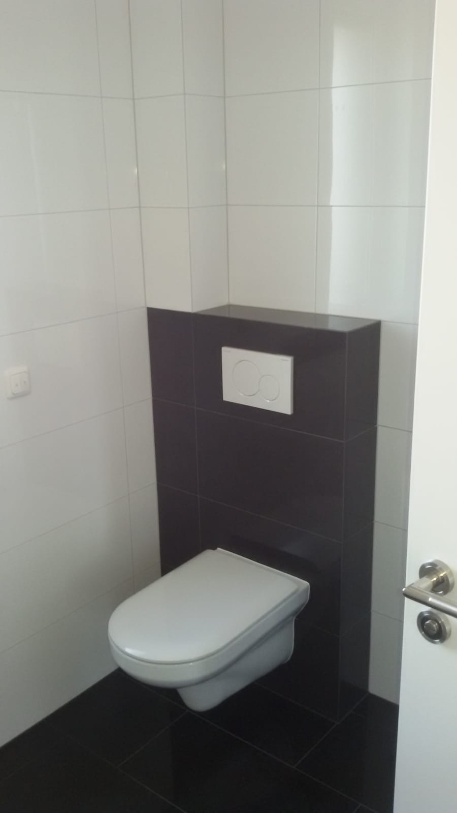 Nowoczesna toaleta podwieszana z białym sedesem i przyciskiem spłukującym na ciemnobrązowej ścianie z płytek, białe płytki na pozostałych ścianach, ciemna podłoga z płytek.