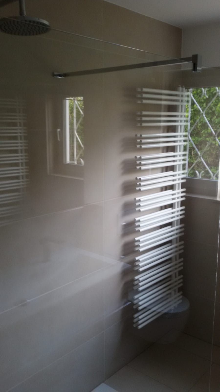 Nowoczesna łazienka z szarymi płytkami, grzejnikiem drabinkowym i deszczownicą sufitową, widok przez szklaną ściankę kabiny prysznicowej.