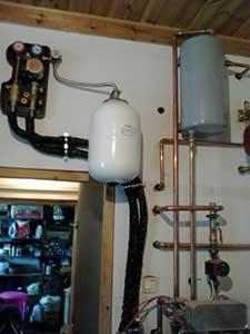 Biały zbiornik wyrównawczy ciśnienia zawieszony na ścianie obok czarnej armatury, z widocznymi miedzianymi rurami i elementami instalacji centralnego ogrzewania, na tle częściowo widocznego...