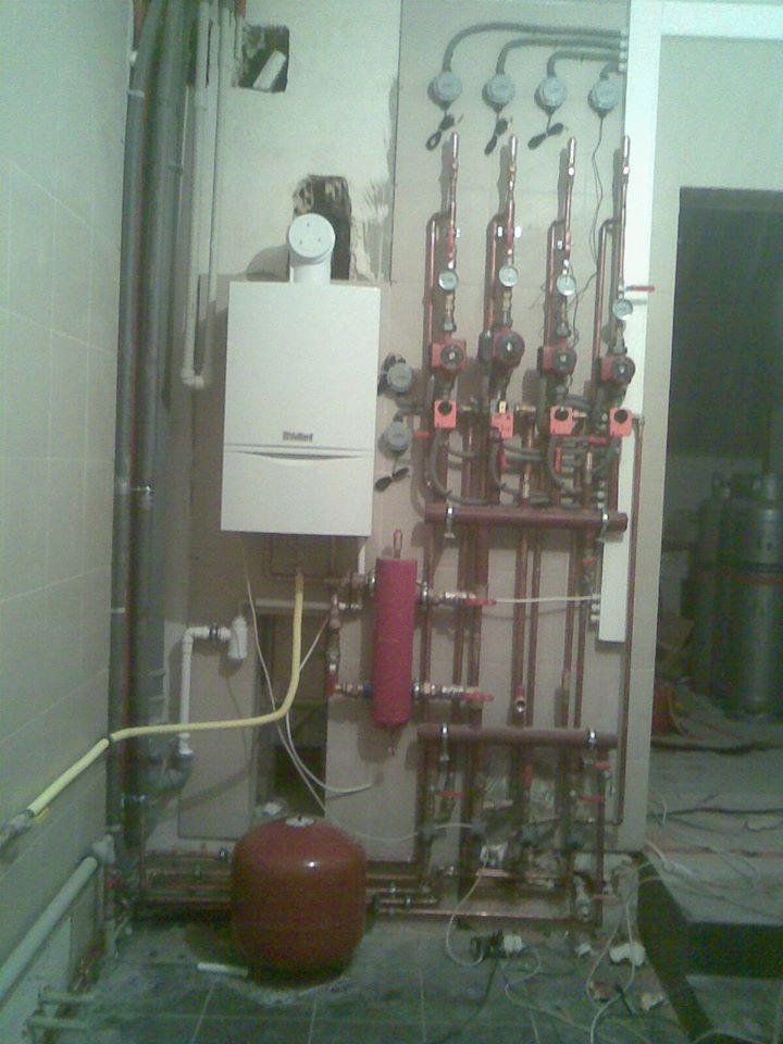 Kotłownia w trakcie instalacji: widoczny kocioł gazowy Vaillant, rozdzielacz z pompami obiegowymi i naczynie przeponowe w kolorze czerwonym. Instalacja w trakcie montażu, widoczne przewody i rury.