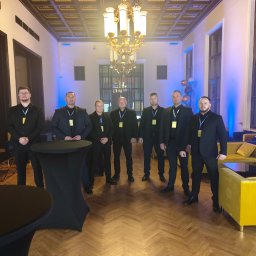 Agencja Ochrony Osób i Mienia Astrea Ochrona Imprez Masowych - Grupa siedmiu ochroniarzy w czarnych garniturach, stojących w eleganckim wnętrzu z parkietem i dekoracjami, gotowych do zabezpieczenia wydarzenia.