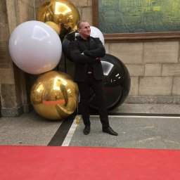 Agencja Ochrony Osób i Mienia Astrea Ochrona Imprez Masowych - Mężczyzna w czarnym garniturze stoi na czerwonym dywanie, otoczony dużymi, błyszczącymi kulami w kolorach złota, czerni i bieli. W tle widoczna kamienna ściana i obraz.