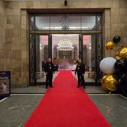 Agencja Ochrony Osób i Mienia Astrea Ochrona Imprez Masowych - Dwóch ochroniarzy w czarnych garniturach stoi na czerwonym dywanie przed wejściem do budynku z dekoracjami balonowymi i żyrandolem.