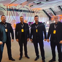 Agencja Ochrony Osób i Mienia Astrea Ochrona Imprez Masowych - Czterech mężczyzn w czarnych garniturach, z identyfikatorami, stoi na tle dekoracji w hali. Uwagę zwraca oświetlenie i dekoracje w tle.