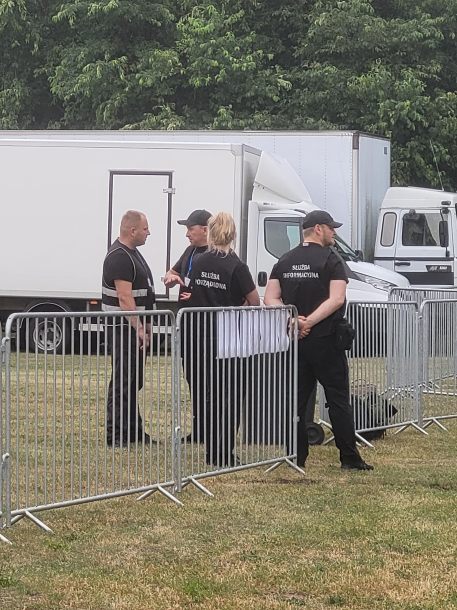 Grupa ochroniarzy w czarnych koszulkach z napisem 'Służba Porządkowa' i 'Służba Informacyjna' stoi na tle białych samochodów dostawczych i metalowego ogrodzenia.
