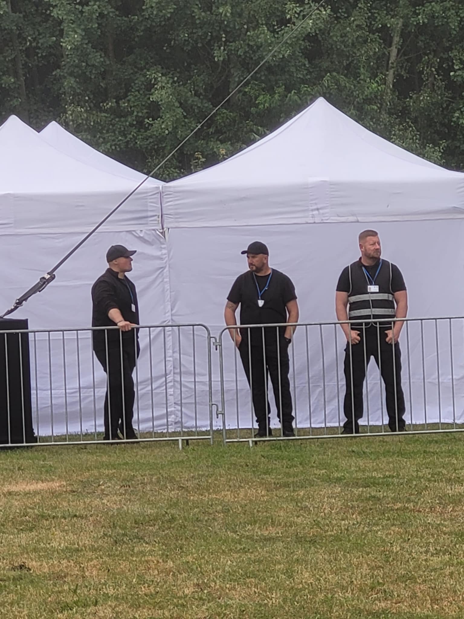 Trzech ochroniarzy w czarnych uniformach za metalowym ogrodzeniem na tle białych namiotów i zieleni.