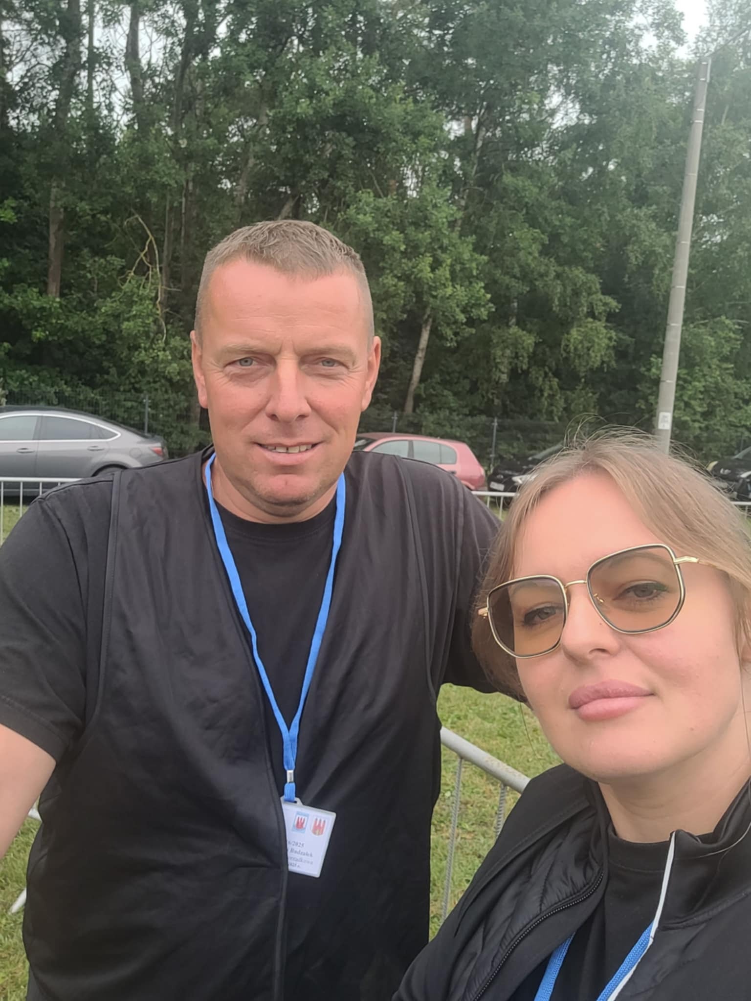 Portret dwóch osób, prawdopodobnie pracowników ochrony, na tle zieleni i zaparkowanych aut. Obie osoby mają identyfikatory na szyi.