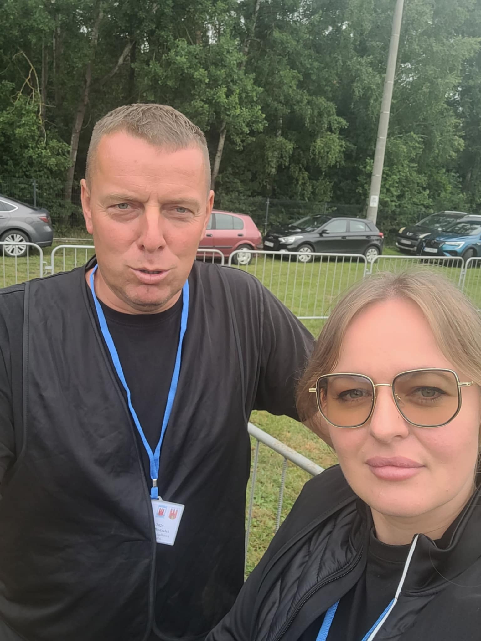 Dwoje pracowników z identyfikatorami na tle parkingu i zieleni. Mężczyzna w czarnej kamizelce, kobieta w okularach. Widoczny fragment barierek.