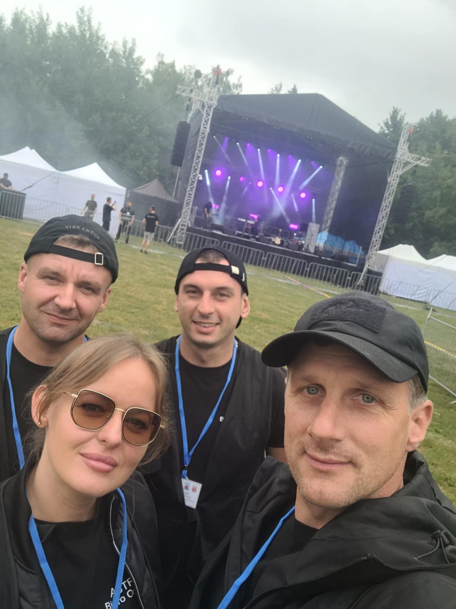 Zespół ochrony na tle sceny koncertowej z reflektorami i białymi namiotami w tle. Mężczyźni w czarnych ubraniach i czapkach, kobieta w okularach.