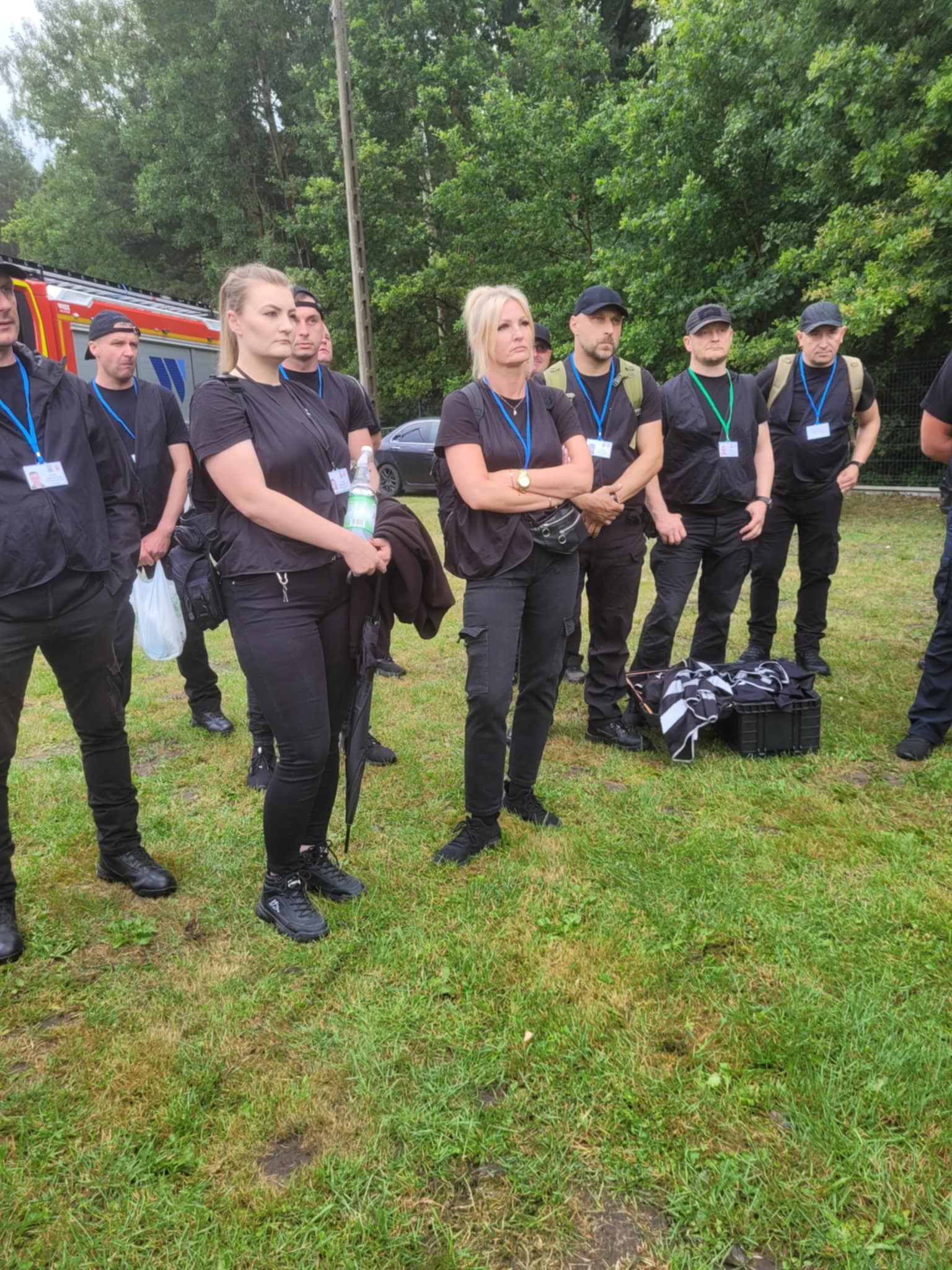 Grupa pracowników w czarnych uniformach z identyfikatorami, stojących na trawie, z wozem strażackim i drzewami w tle. Obok nich leży czarno-biały ręcznik.